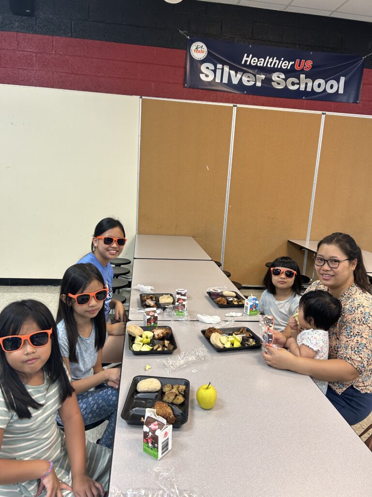 A photo of four children and a baby on a woman's lap, eating SUN Meals in a school cafeteria.