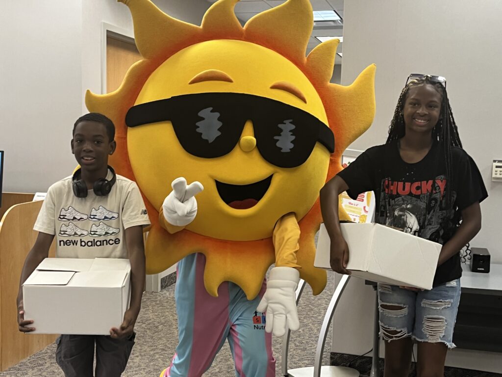 A photo of NC Summer Meals mascot, Ray F. Sunshine, with two children holding Meals To-Go boxes of food.