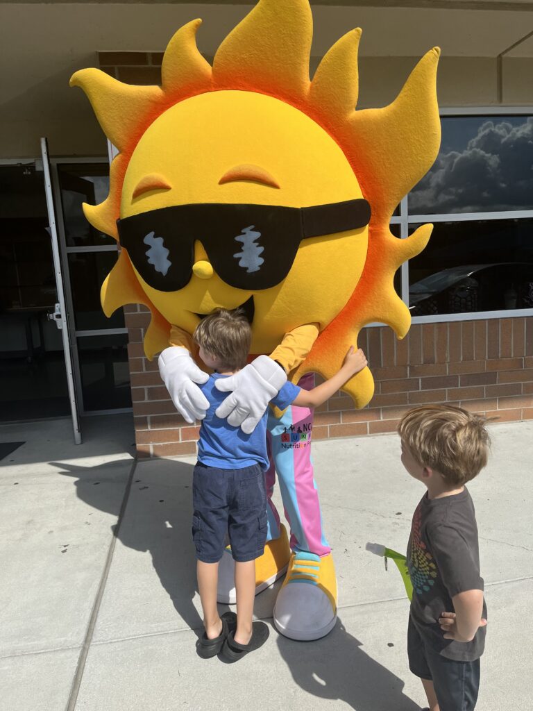 A ;photo of NC Summer Meals mascot Ray F. Sunshine, hugging a child and another child watches nearby.